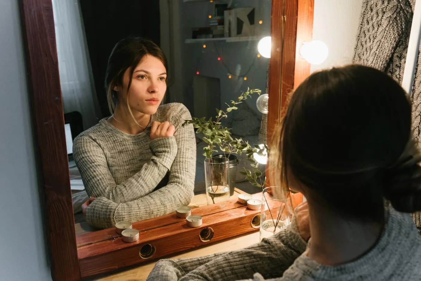Femme en pull qui contemple son reflet dans un miroir, dans une pièce à la lumière chaleureuse